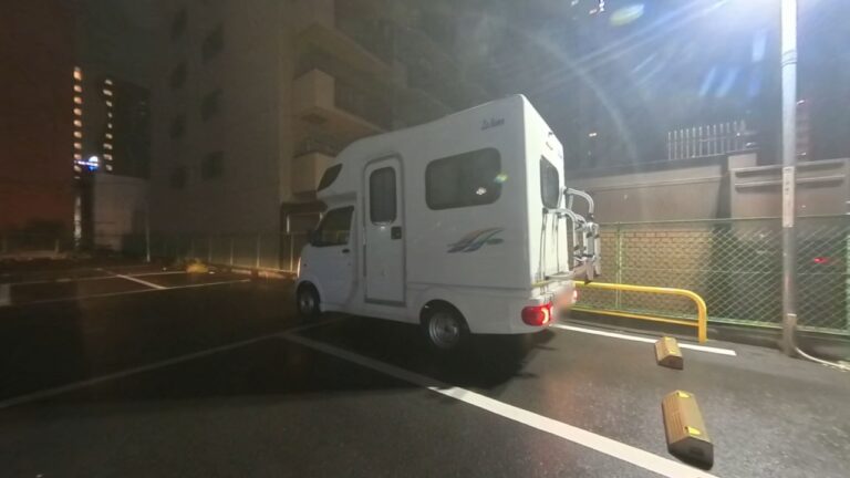 雨に濡れたコインパーキングに停めた白い軽キャンピングカー・ラクーン